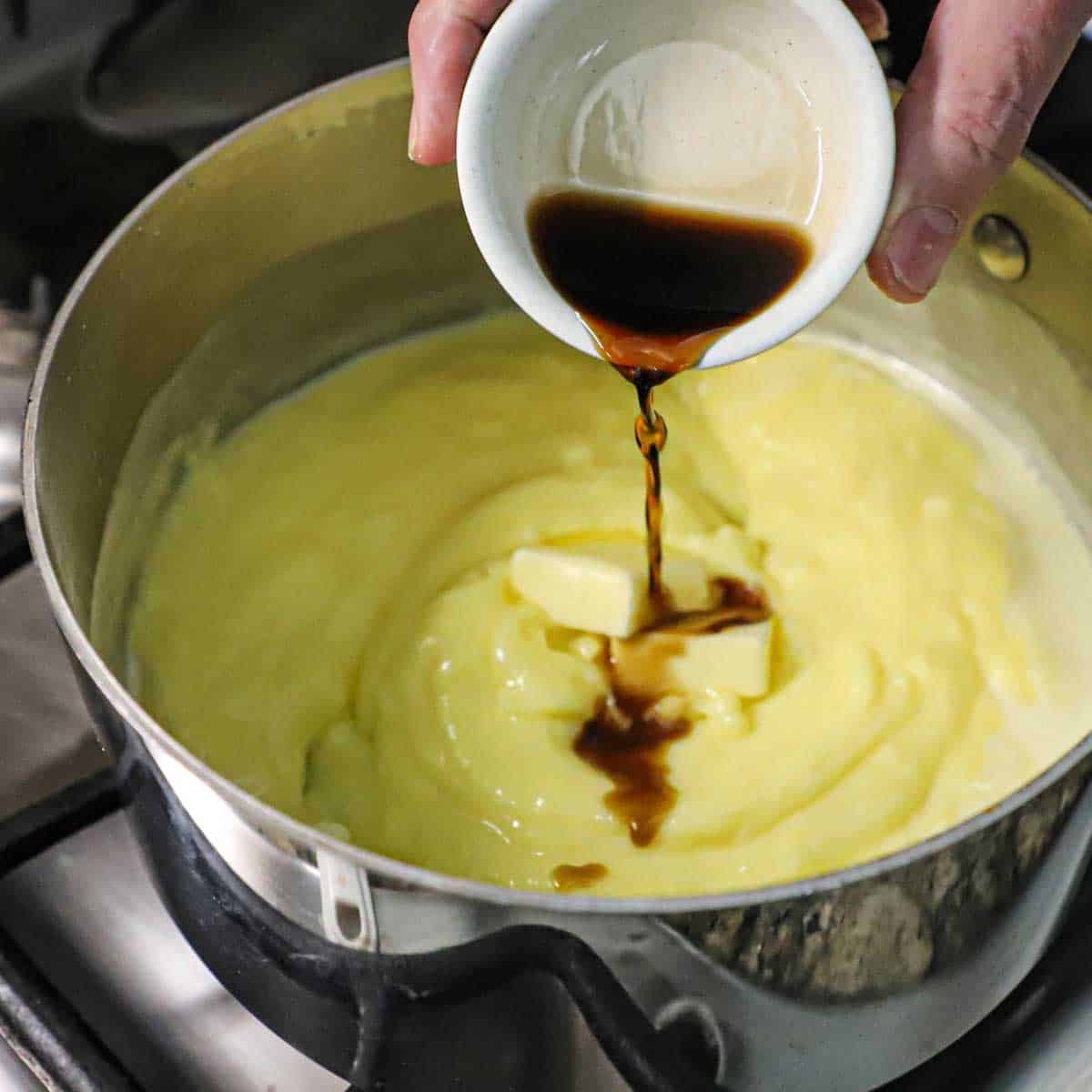 A person pouring vanilla extract from a small bowl into a saucepan that is filled with a vanilla custard.
