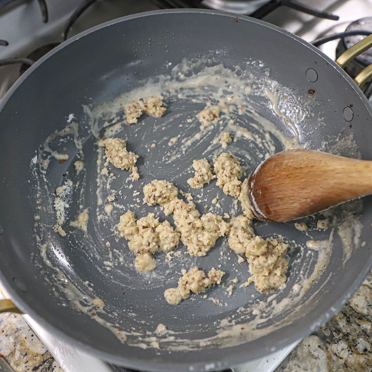 A person using a large wooden spoon to stir flour combined with oil and melted butter to create a roux in a large non-stick skillet on a gas stove.