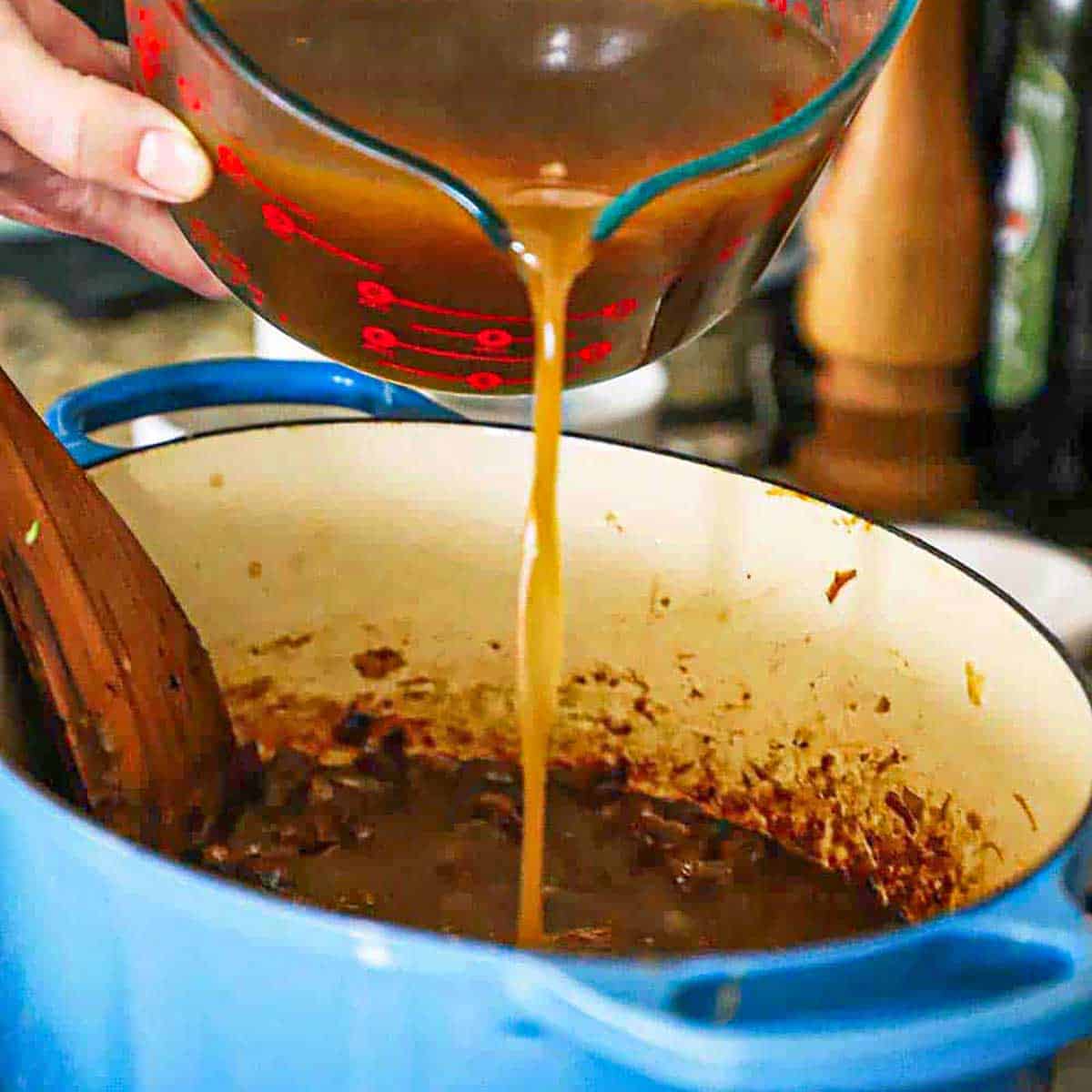 A person pouring homemade beef stock from a large glass measuring cup into a Dutch oven that is filled with deeply caramelized onions.