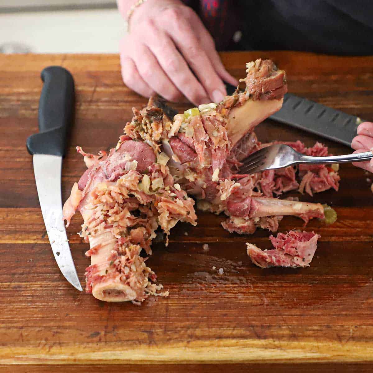A person using a fork to pull cooked ham from a large ham bone that is resting on a wooden cutting board with two chef's knife resting nearby.