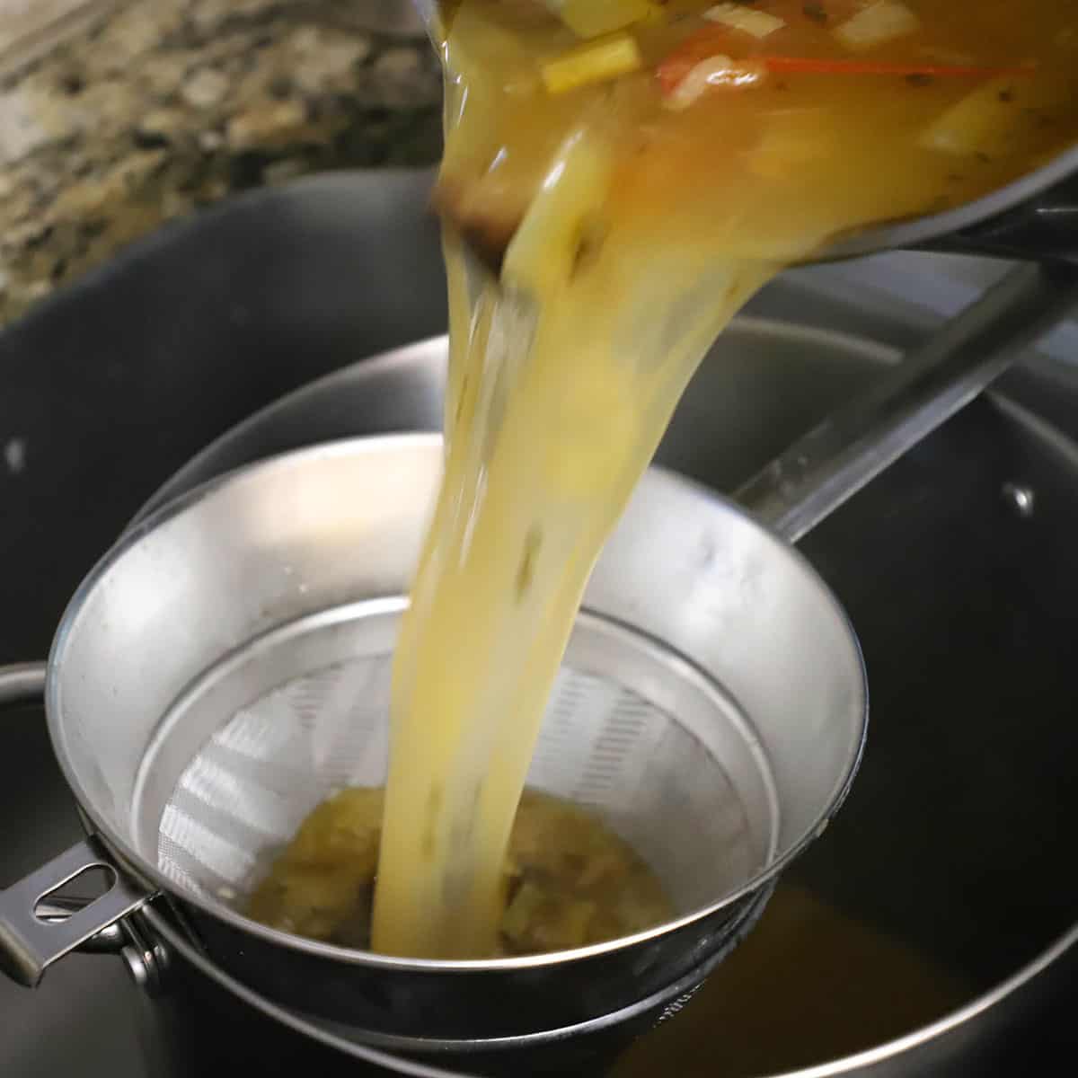 A person straining homemade beef stock from a stock pot into another stock pot and a fine-mesh sieve.