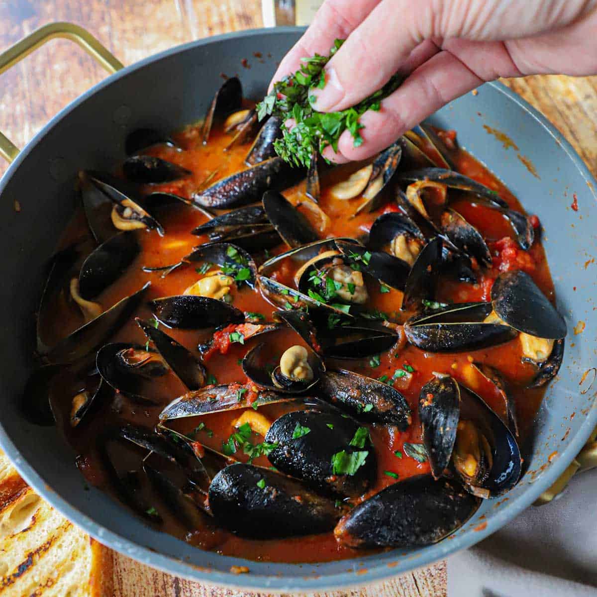 A person sprinkling freshly chopped basil and parsley onto a skillet filled with freshly prepared Easy Mussels Marinara.