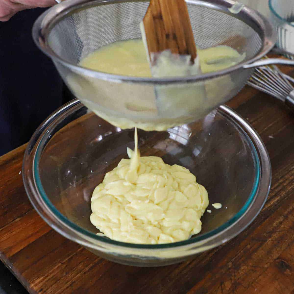 A person using a wooden spatula to pass a vanilla custard through a fine-mesh sieve into a glass bowl sitting on a wooden cutting board.