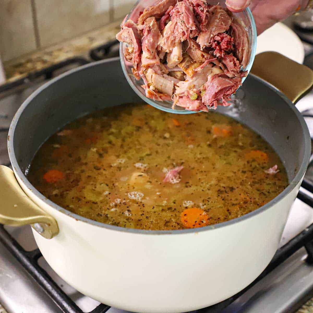 A person transferring chopped cooked ham from a glass bowl into a stock pot filled with simmering classic ham and bean soup on a gas stove.