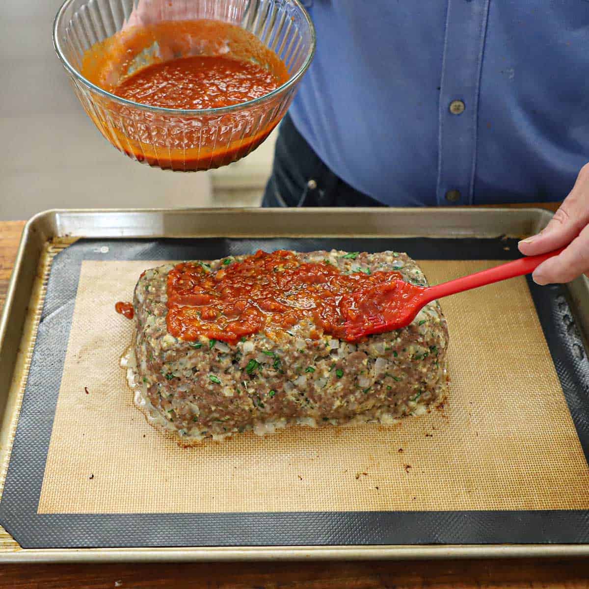 A person using a red pastry brush to apply a marinara and balsamic glaze onto a partially cooked Italian-style meatloaf on a baking sheet.