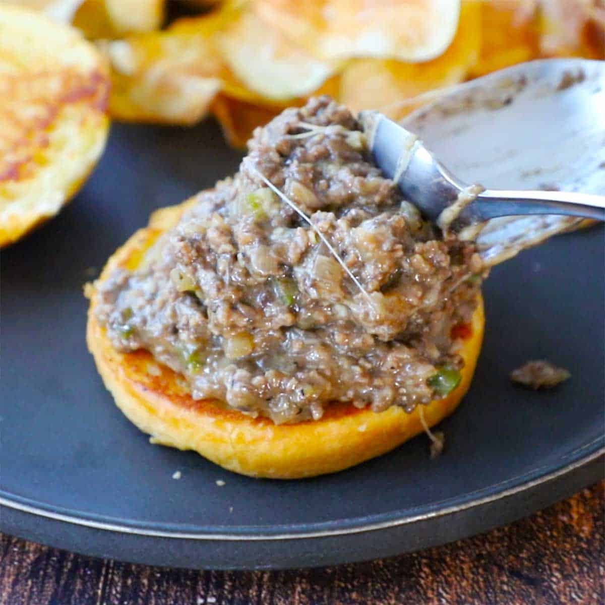 A person using a spoon to place a pile of Philly cheesesteak sloppy Joes onto the bottom of a toasted Brioche bun on a black plate.