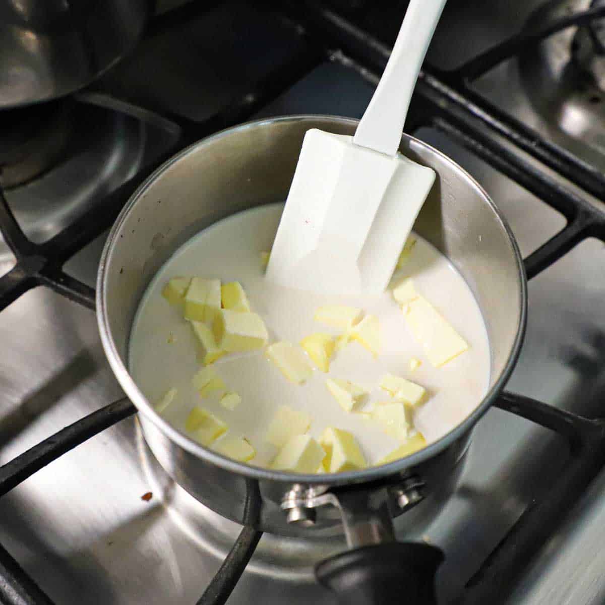 A person using a white rubber spatula to stir cubed butter and simmering milk in a small saucepan on a gas stove.