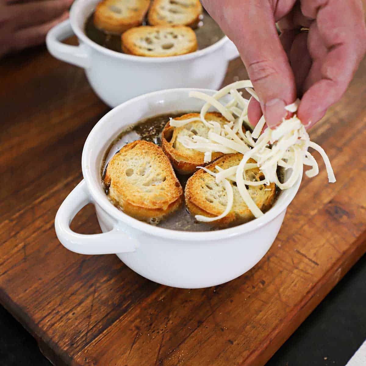 A person placing shredded Gruyere cheese over the top of three toasted baguette slices that are resting on the surface of a bowl of homemade French onion soup.