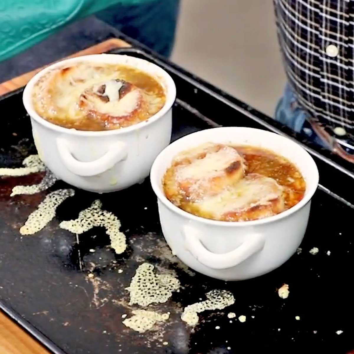 A person wearing oven mitt holding a baking sheet that has two soup bowls that are filled with homemade French onion soup that have just been under the broiler to melt the cheese.