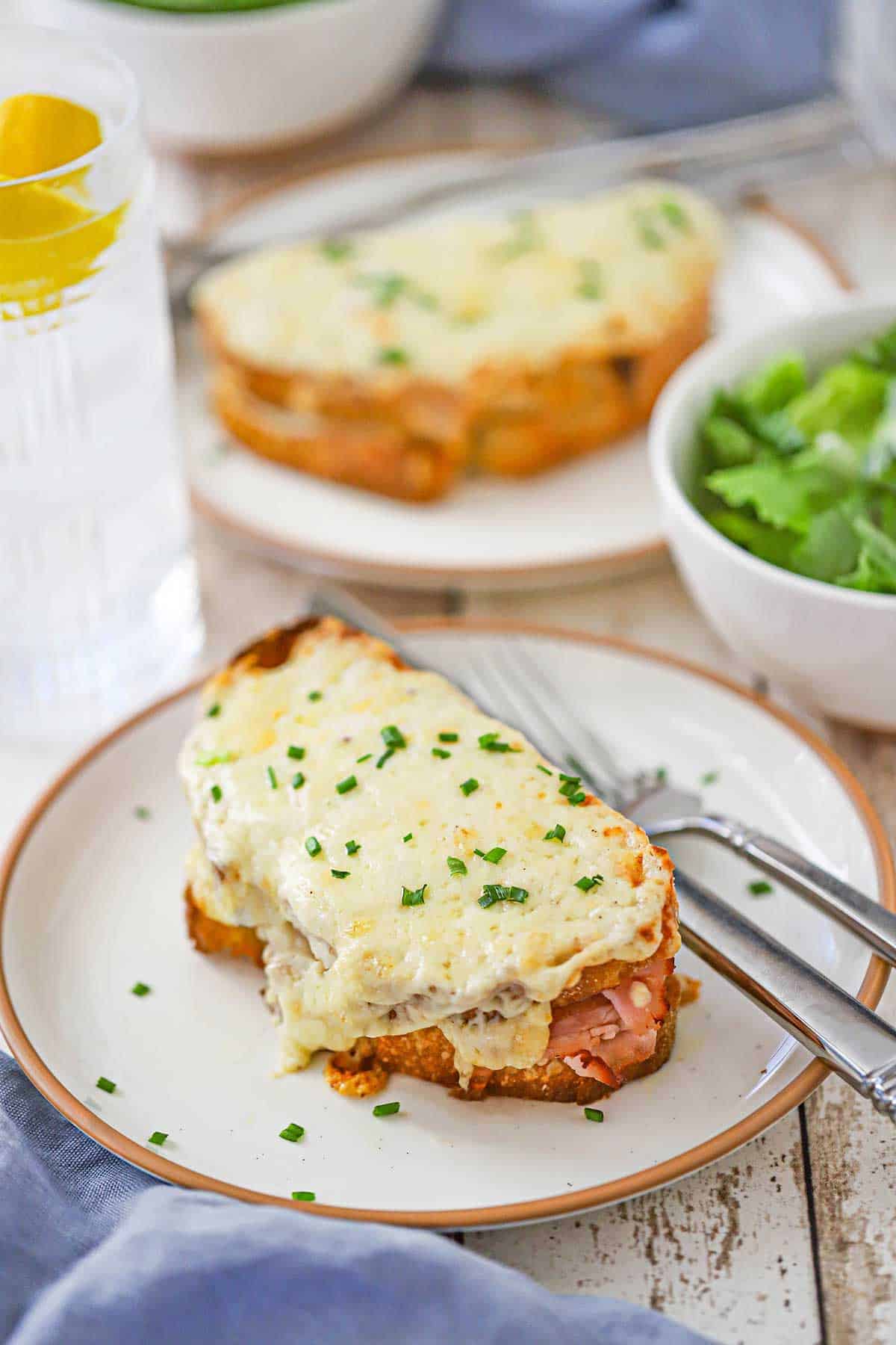 An overhead view of two plates both filled with a freshly prepared Essential Croque Monsieur with a bowl filled with a green salad nearby.