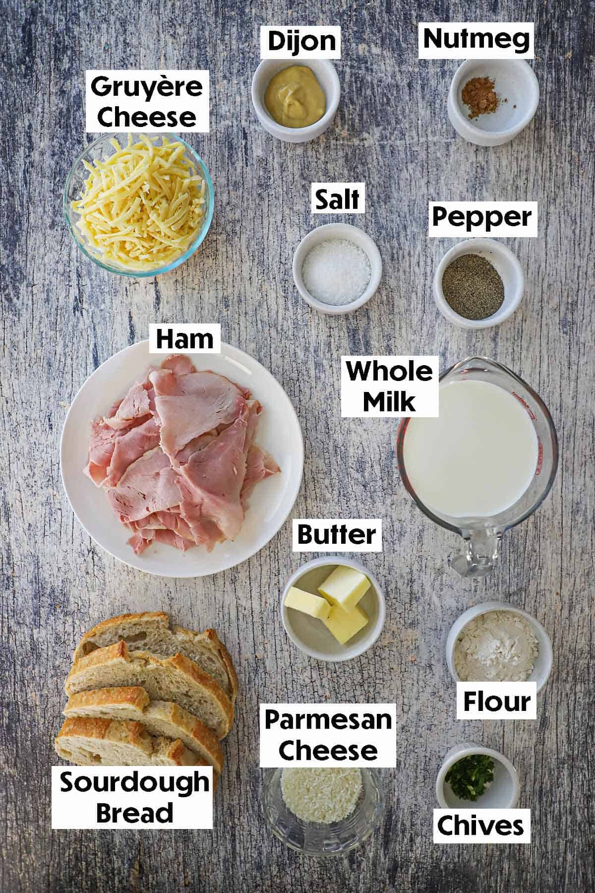 An arrangement of ingredients for The Essential Croque Monsieur on a grey wooden background including Gruyere cheese, sliced ham, sourdough bread slices, Dijon mustard, milk butter, flour, Parmesan, and seasonings.
