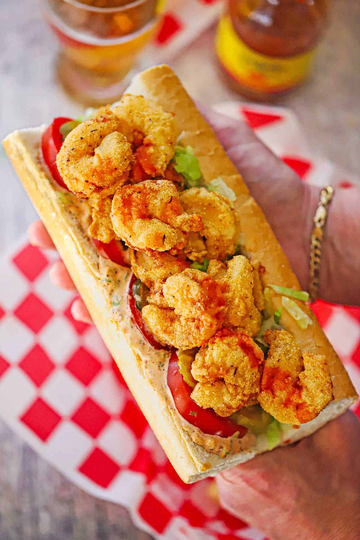 A person using two hands to hold a large New Orleans Fried Shrimp Po-boy over a basket lined with red and white checkered wax paper.