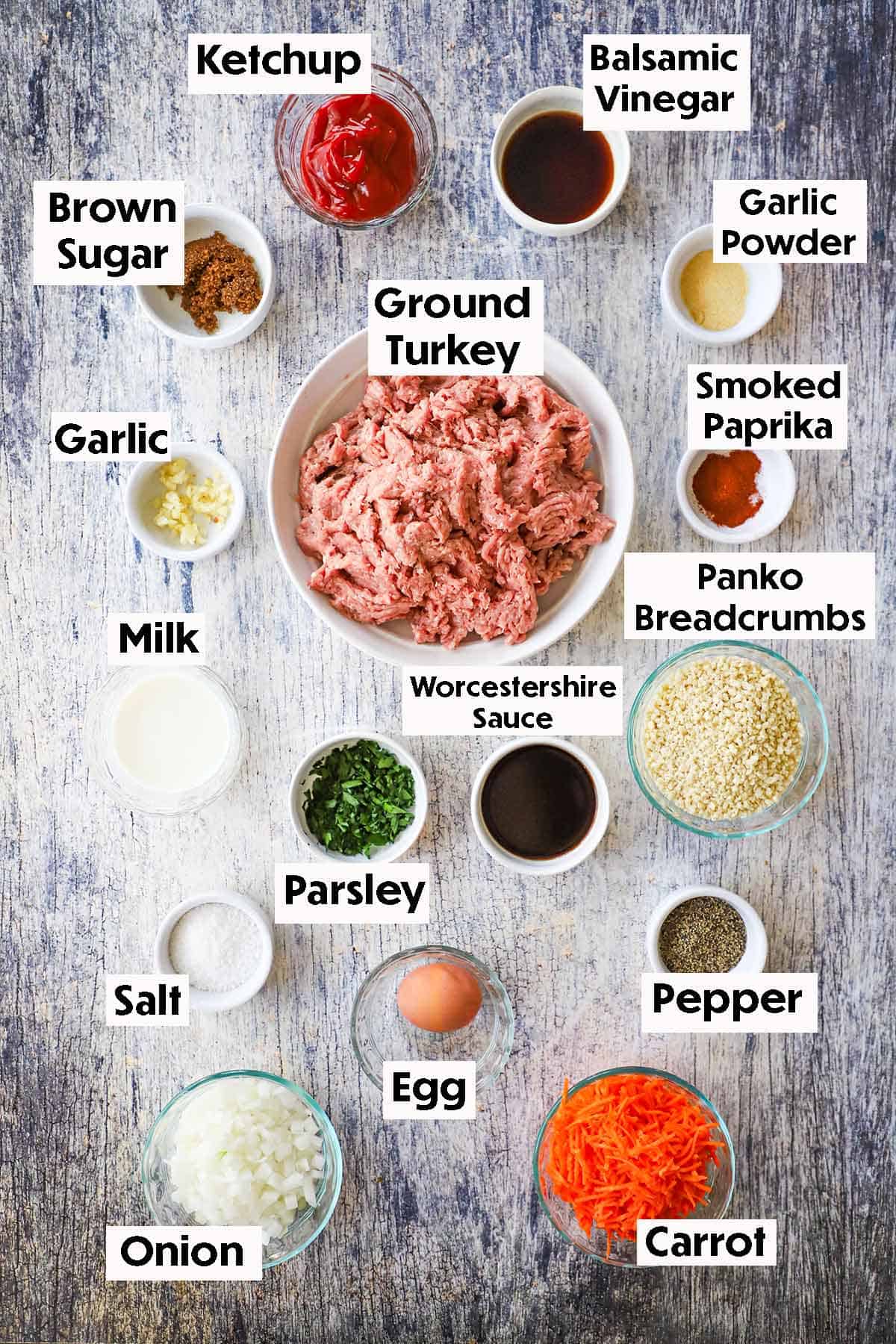 An arrangement of ingredients on a grey wooden background including ground turkey, breadcrumbs, shredded carrots, onions, parsley, garlic, milk, brown sugar, ketchup, vinegar, and other seasonings.
