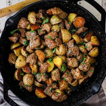 An overhead view of Garlic Butter Steak Bites and Potatoes in a large black cast-iron skillet garnished with chopped Italian parsley and coarse sea salt.