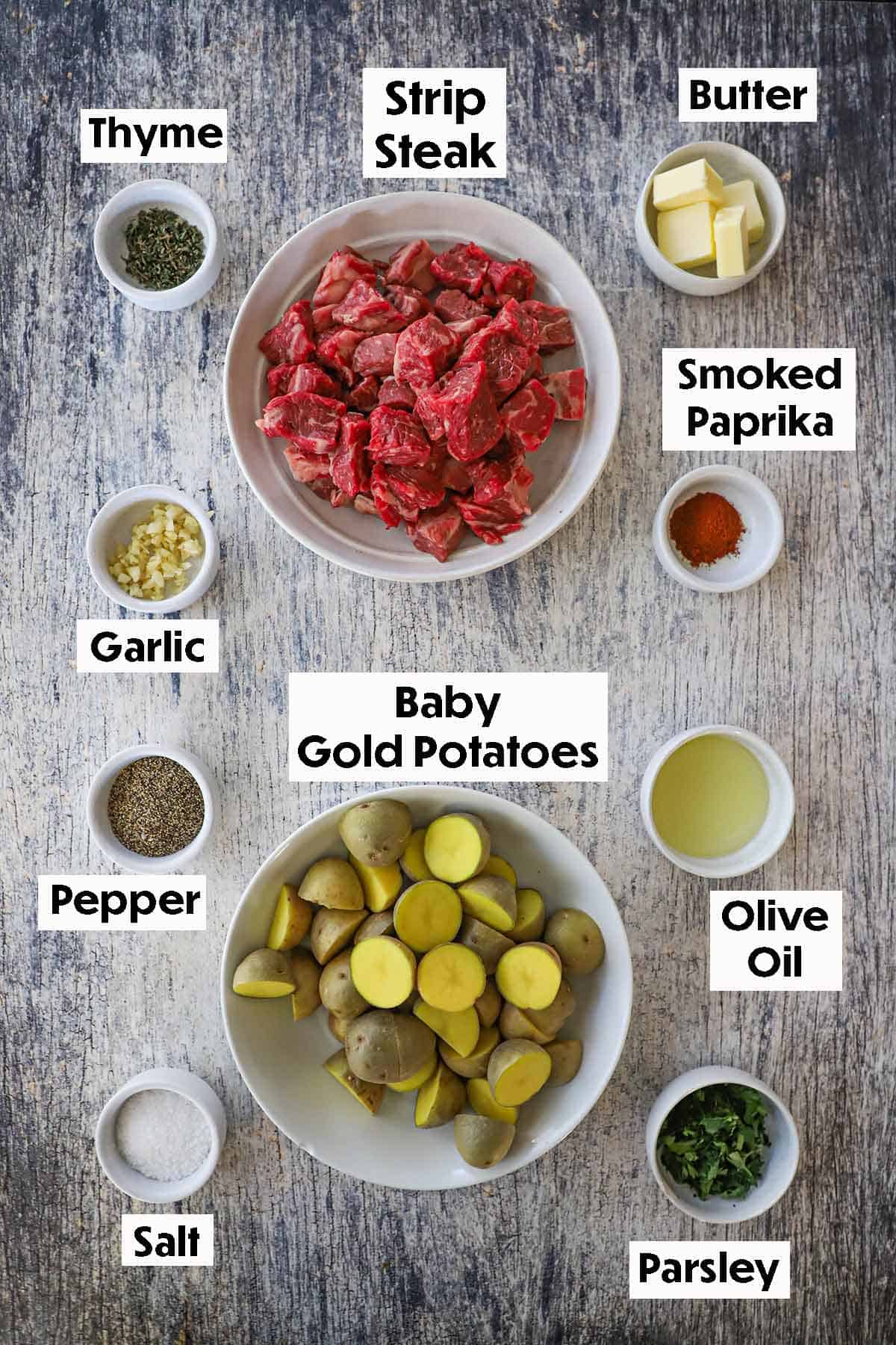 An arrangement of ingredients for Garlic Butter Steak Bites and Potatoes on a grey wooden background including cubed steak, baby gold potatoes, garlic, butter, olive oil, and seasonings.