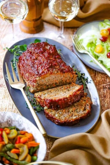 A juicy turkey meatloaf that has two slices cut away resting on an oval blue platter with gold serving fork nearby, as well as two glasses of white wine and a green salad.