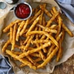 An overhead view of a pile of Crispy Beer Battered French Fries on a crumpled piece of brown paper with a small vessel filled with ketchup nearby.
