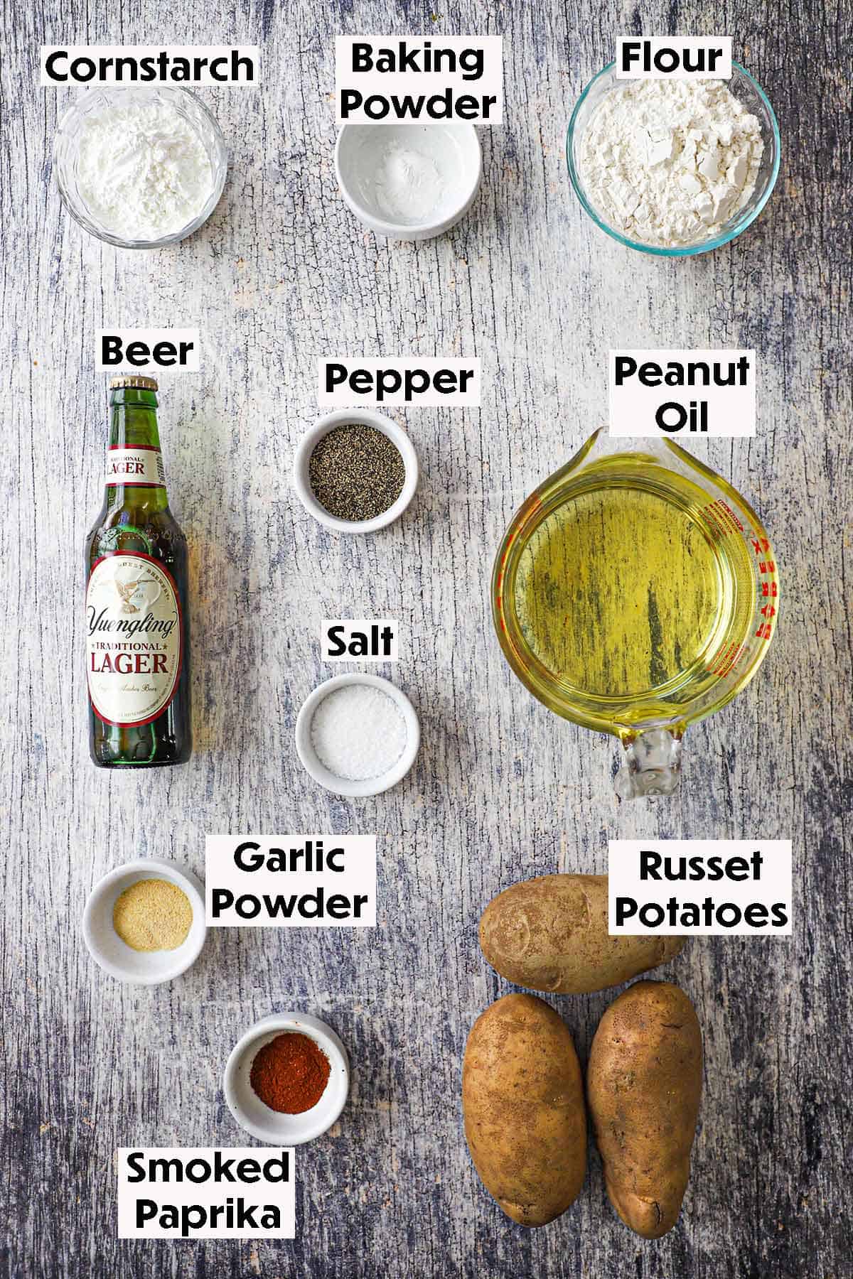 An arrangement of ingredients for Crispy Beer Battered French Fries on a grey wooden background including Russet potatoes, a bottle of better, conrstarch, baking powder, flour, oil, and seasonings.