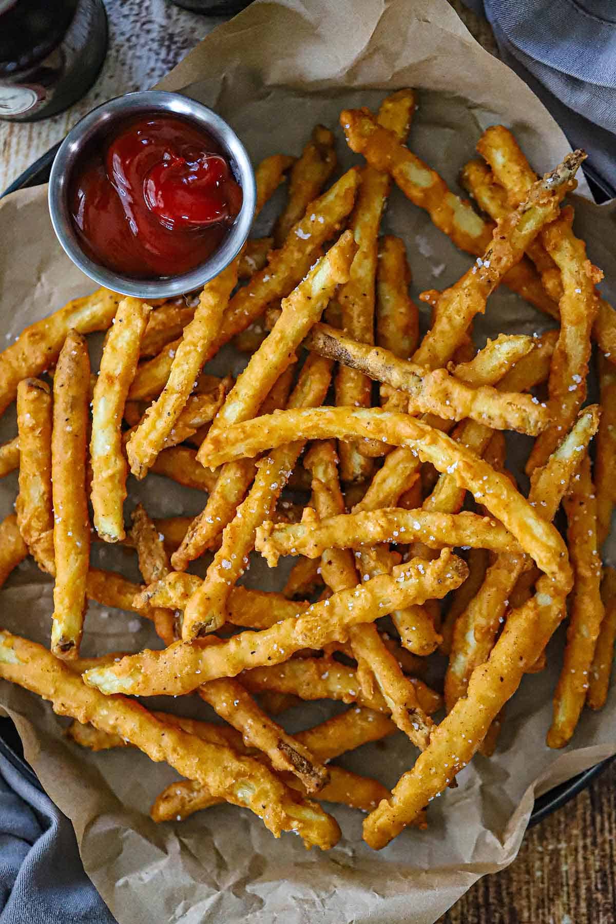 An overhead view of a pile of Crispy Beer Battered French Fries on a crumpled piece of brown paper with a small vessel filled with ketchup nearby.