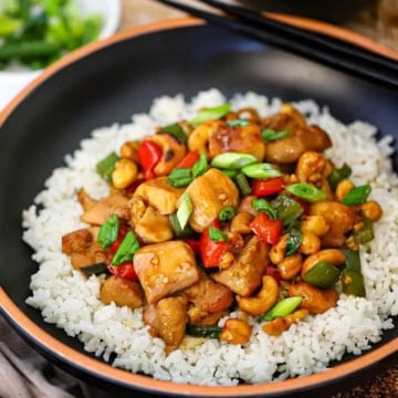 A close-up view of a serving of Cashew Chicken resting on a bed of white rice in a black bowl with chopsticks resting on the side.