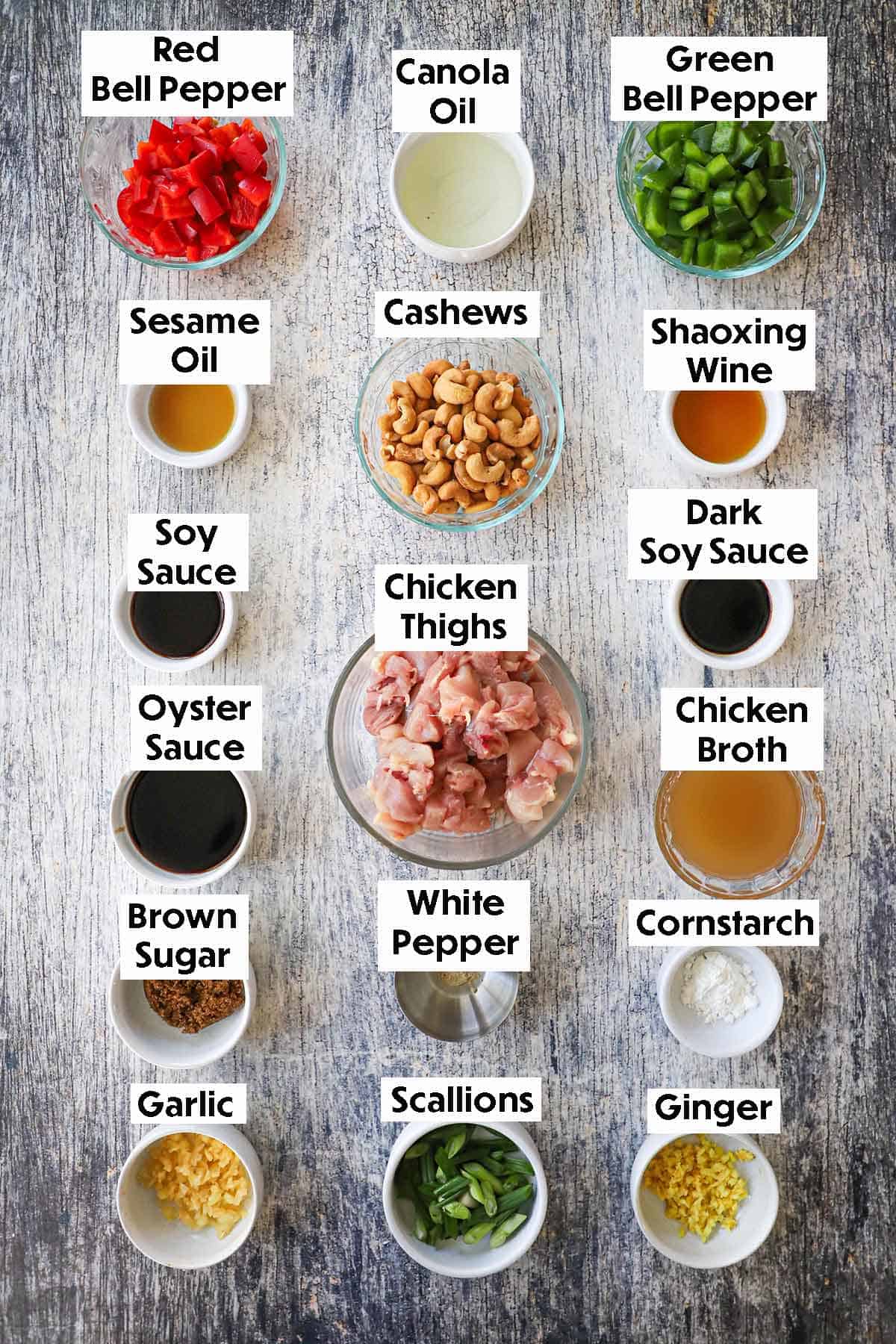 An arrangement of ingredients for cashew chicken on a grey wooden background including chicken, cashews, bell peppers, garlic, ginger, chicken broth, scallions, and Chinese flavor boosters.
