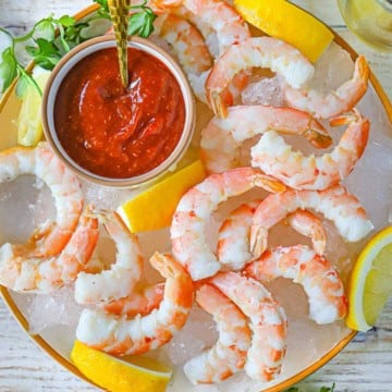 An overhead view of Classic Jumbo Shrimp Cocktail on a white plate lined with crushed ice with lemon wedges placed throughout and a small bowl filled with homemade cocktail sauce off to the side of the shrimp.