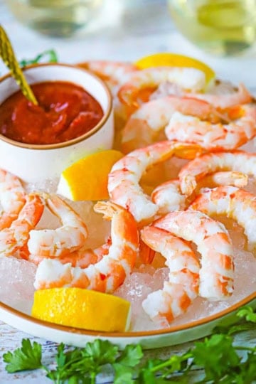 A close-up view of Classic Jumbo Shrimp Cocktail on a plate filled with shaved ice, lemon wedges and a small bowl filled with homemade cocktail sauce.