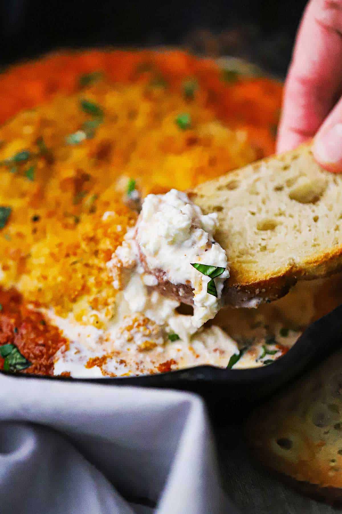 A person using a thin piece of toast to lift creamy burrata from a cast-iron skillet the is filled with a crispy fried burrata with smokey romesco sauce.