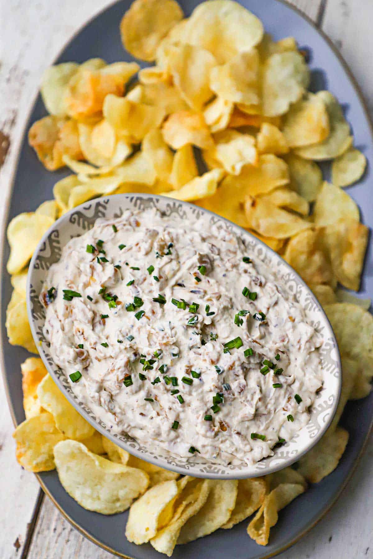 An overhead view of an oval bowl filled with homemade French onion dip garnished with snipped chives on a platter filled with Kettle potato chips.