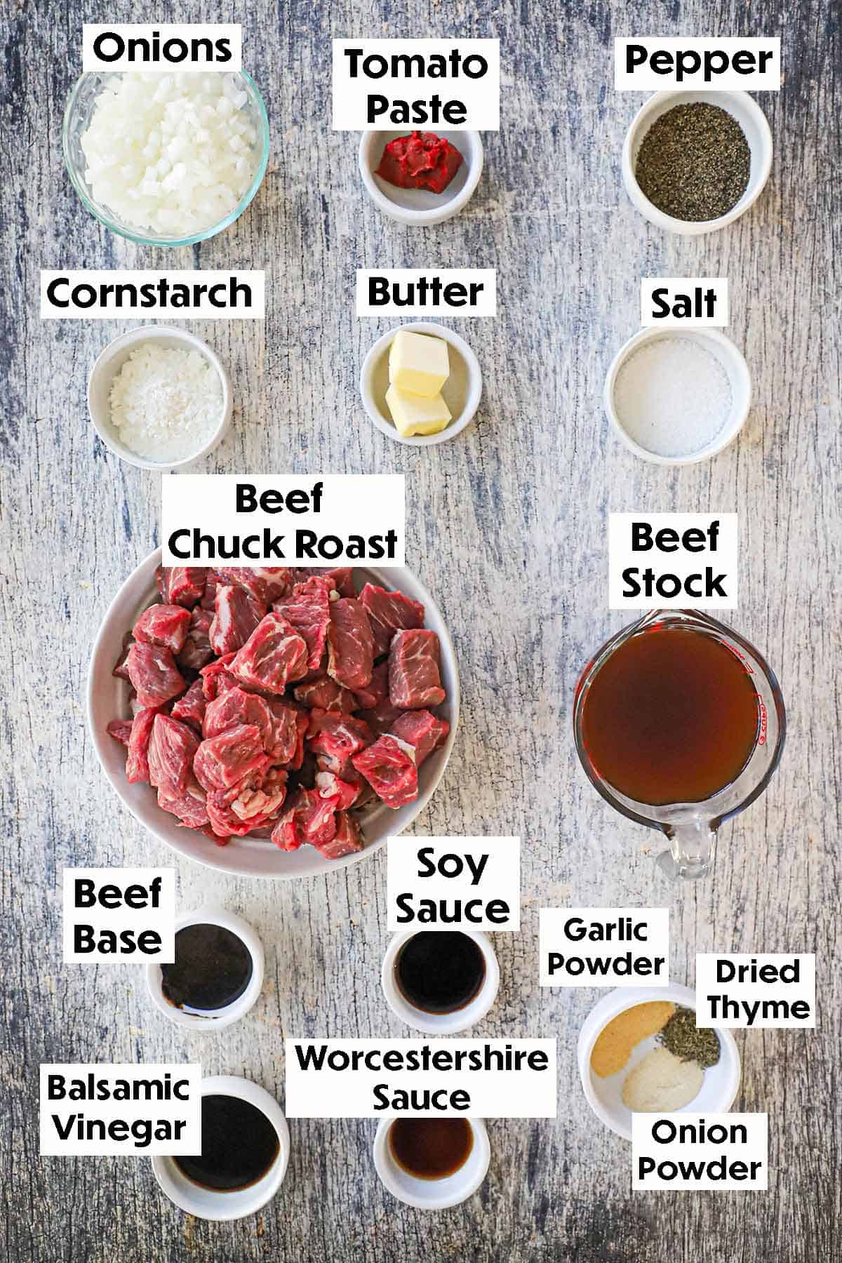 An arrangement of ingredients of slow cooker beef tips and gravy on a grey wooden background including chunks of beef chuck roast, chopped onions, tomato paste, cornstarch, beef stock, soy sauce, Worcestershire sauce, balsamic vinegar, and seasonings.