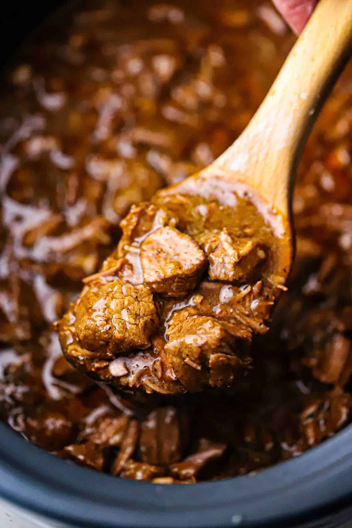 A close up view of a large wooden spoon being used to raise a helping of slow cooker beef tips and gravy from the slow cooker holding the dish.