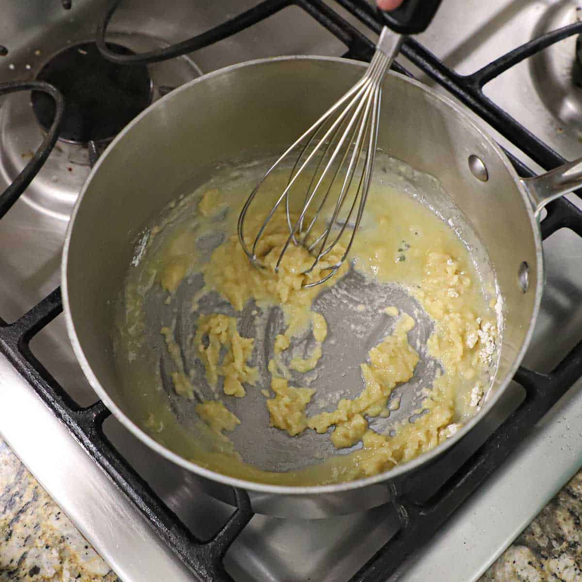 A person using a whisk to create a roux of melted butter and all-purpose flour in the bottom of a steel saucepan on a gas stove.