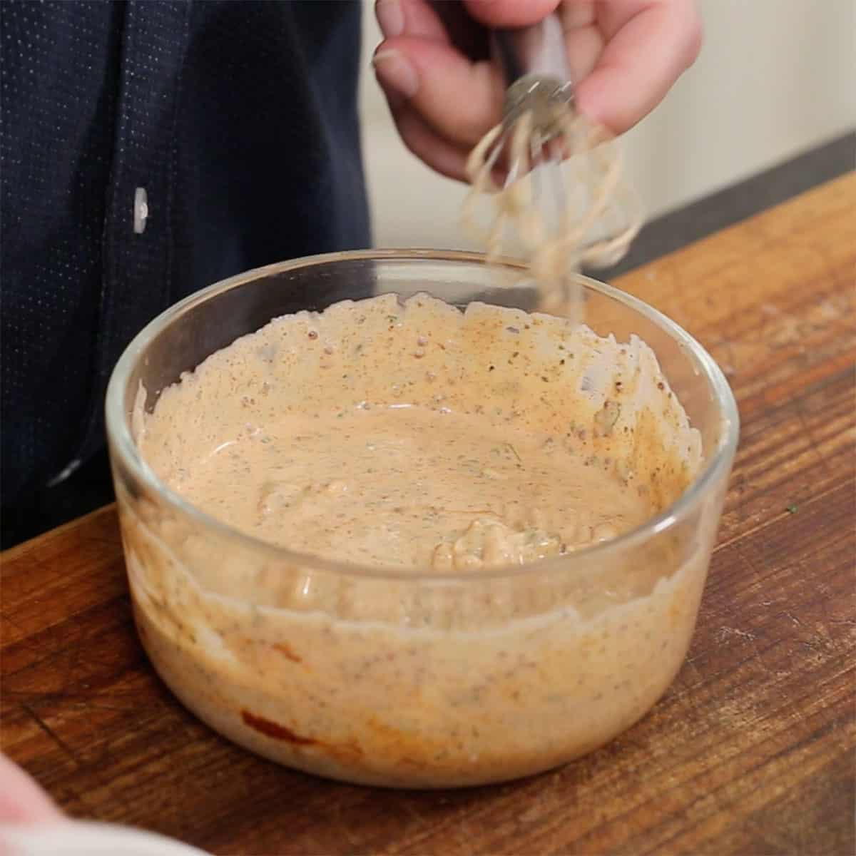 A person using a whisk to combine the ingredients for Cajun Remoulade in a glass bowl on a wooden cutting board.