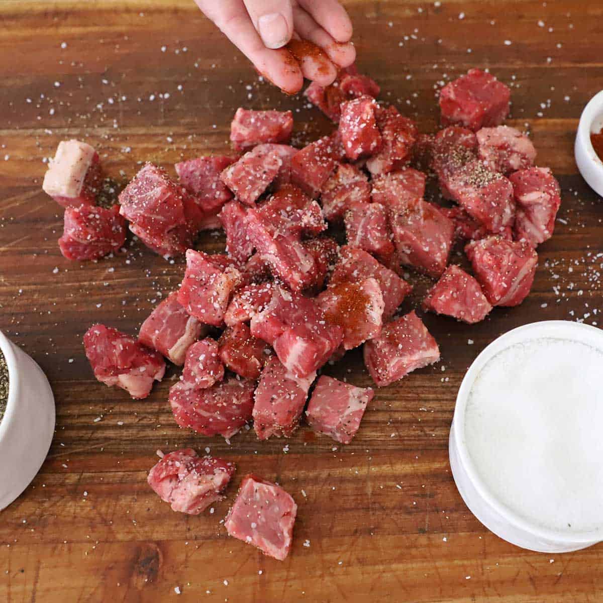 A person sprinkling smoked paprika over a pile of cube strip steak that has also been seasoned with salt and pepper on a wooden cutting board.