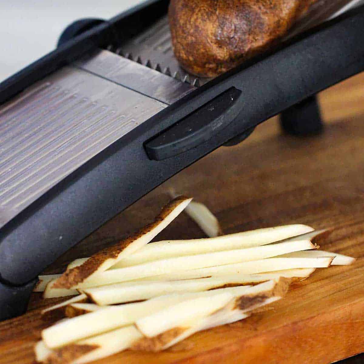 A person using a mandolin to slice a large Russet potato into long strips of potato to be turned into French fries.