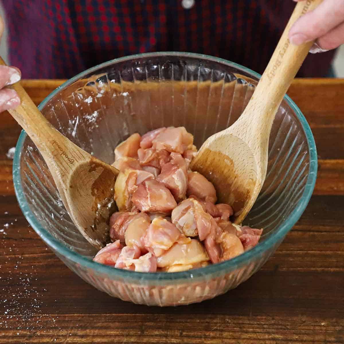 A person using two wooden spoons to toss together cubed chicken thighs with a Chinese marinade in a glass bowl on a wooden cutting board.