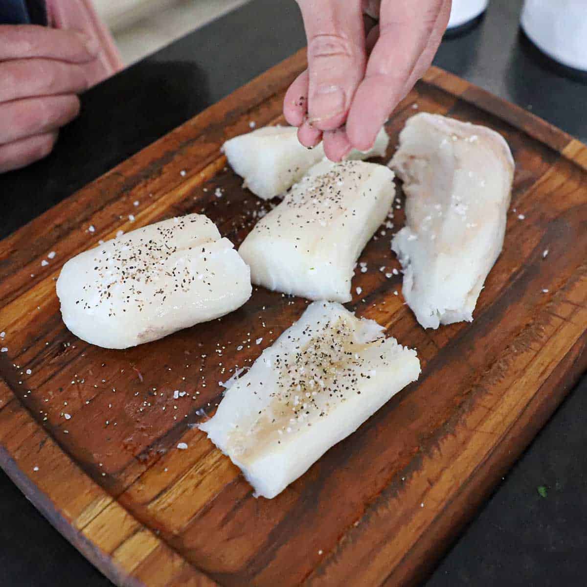 A person using his fingers to sprinkle salt and pepper all over uncooked filets of cod on a small wooden cutting board.