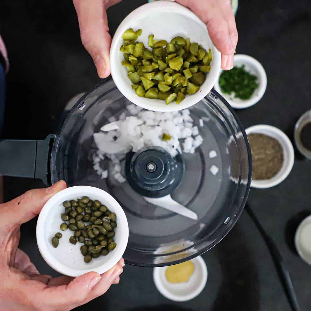A person holding a small bowl of chopped pickles in one hand and a small bowl of capers in the other hand both over a food processor with chopped onions in the bottom of it.