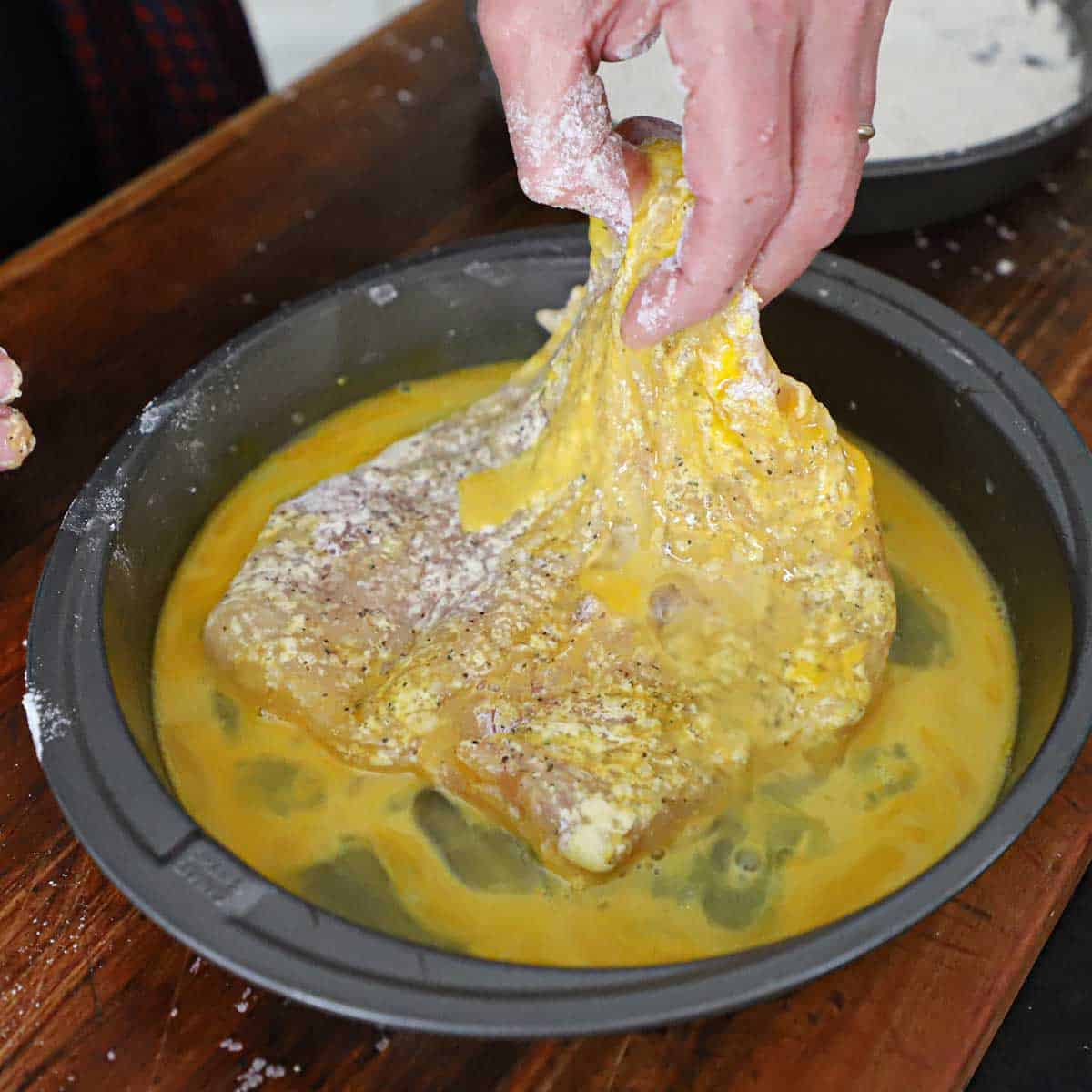 A person dredging a thin chicken cutlet in an egg bath in a shallow baking pan.