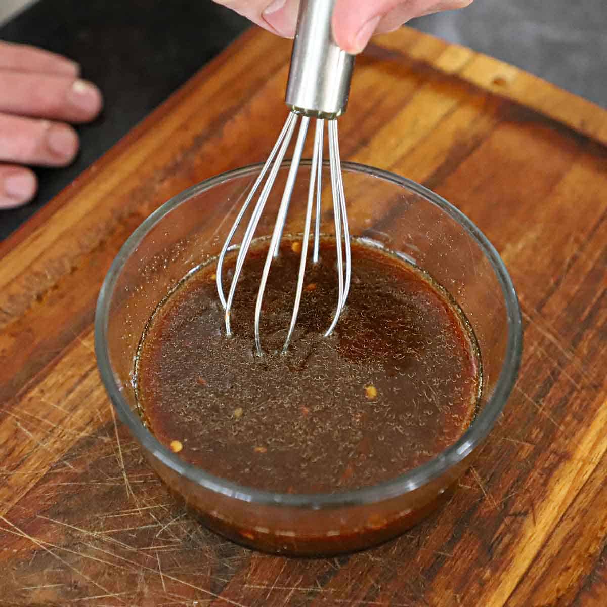 A person whisking an Asian dipping sauce in a glass bowl on a wooden cutting board.