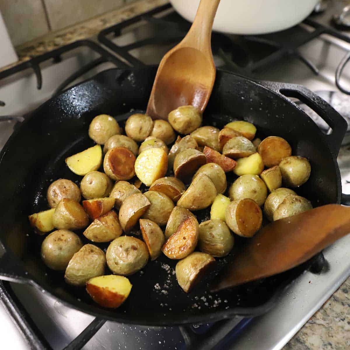 A person using two wooden utensils to stir baby gold potatoes that have been halved and are being seared in a large cast-iron skillet.