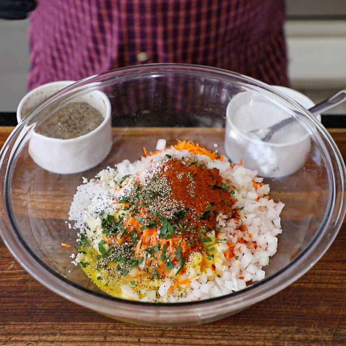 A person standing behind a glass bowl on a wooden cutting board filled with chopped onions, carrots, egg, parsley, and seasonings.