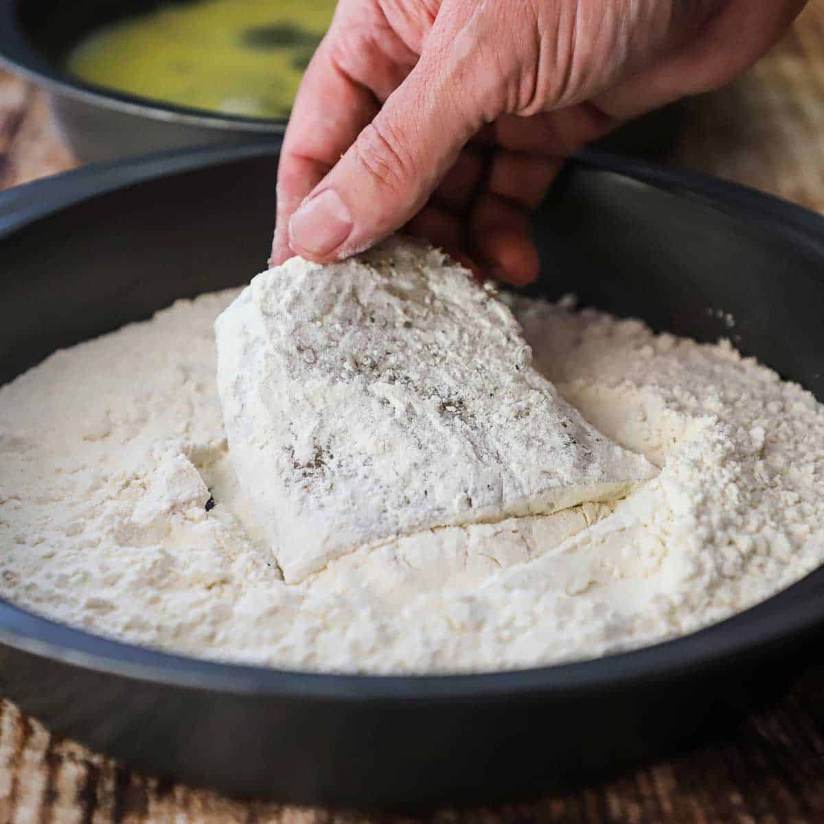 A person dredging a seasoned filet of cod through a pan filled with all-purpose flour.