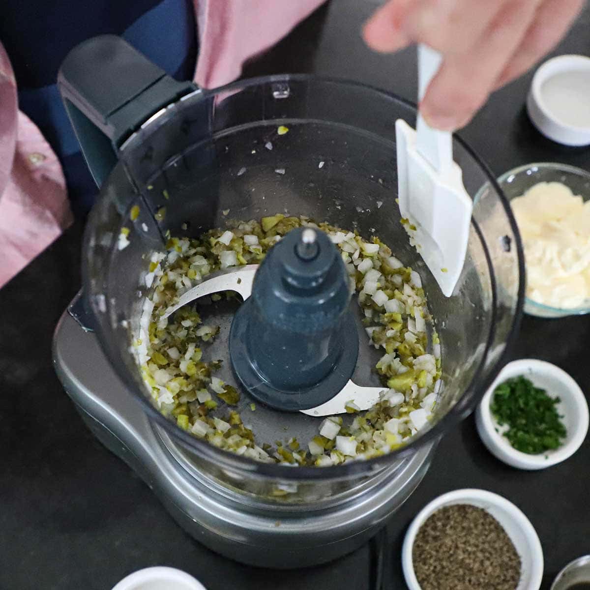 A person using a white rubber spatula to scrape down the sides of a food processor bowl that has chopped pickles, onions, and capers in it.