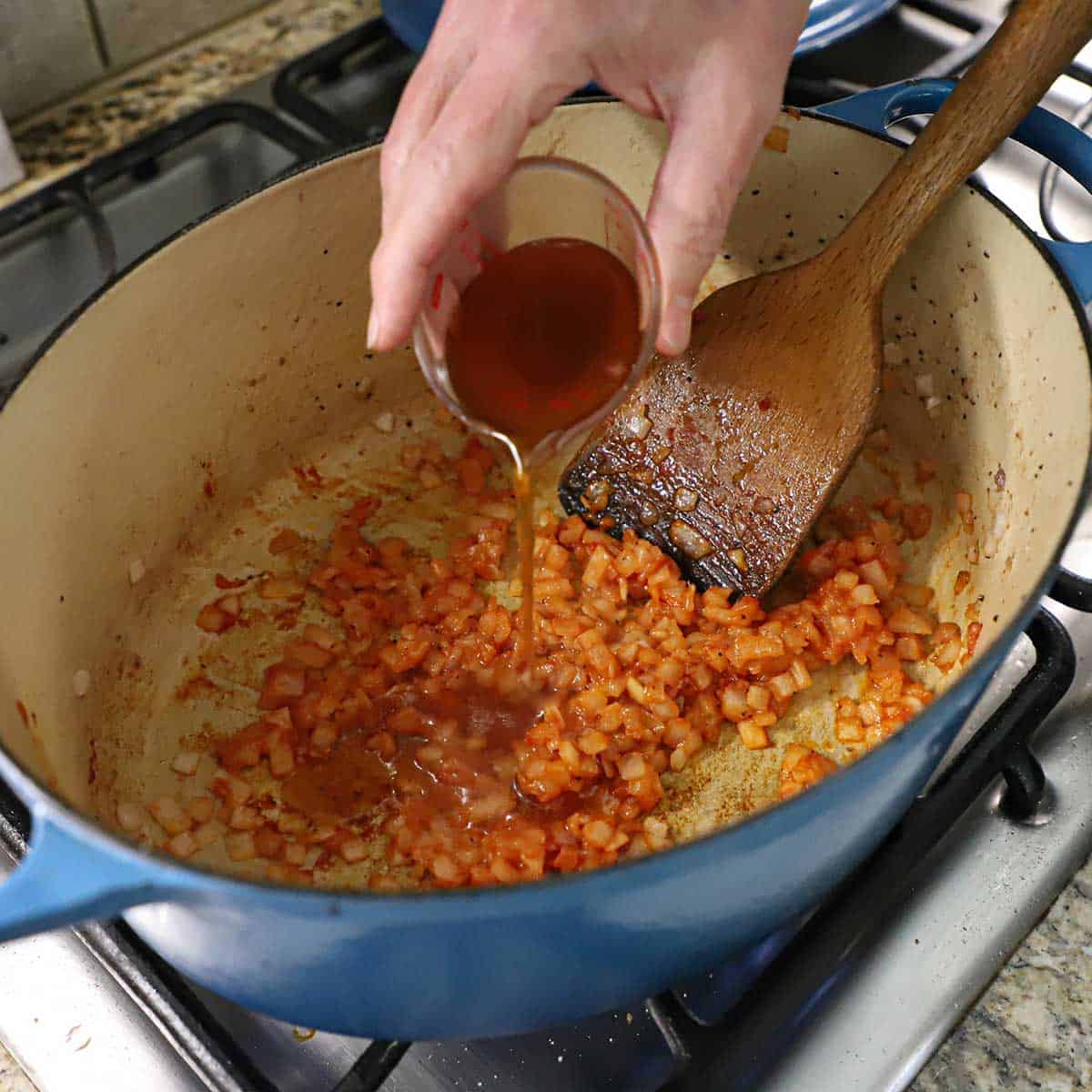 A person pouring a quarter cup of beef stock from a small container into a large Dutch oven that has onions that have been sautéd in oil and tomato paste.