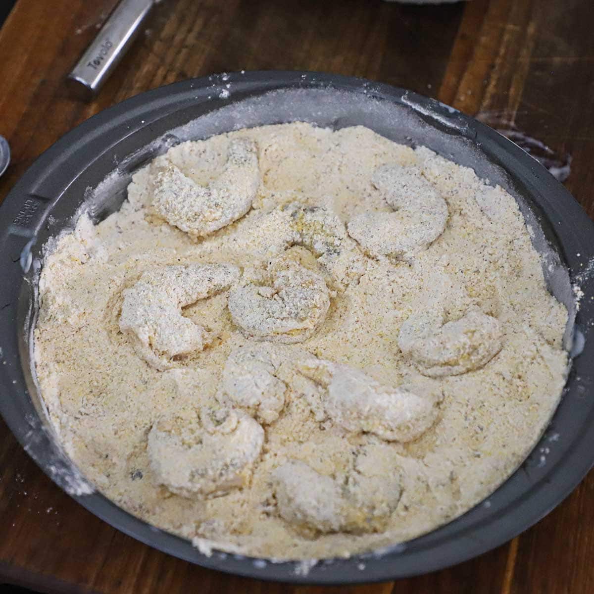 A round baking pan that is filled with a breading mixture of cornmeal, flour, and seasonings with fully breaded shrimp resting in the pan.