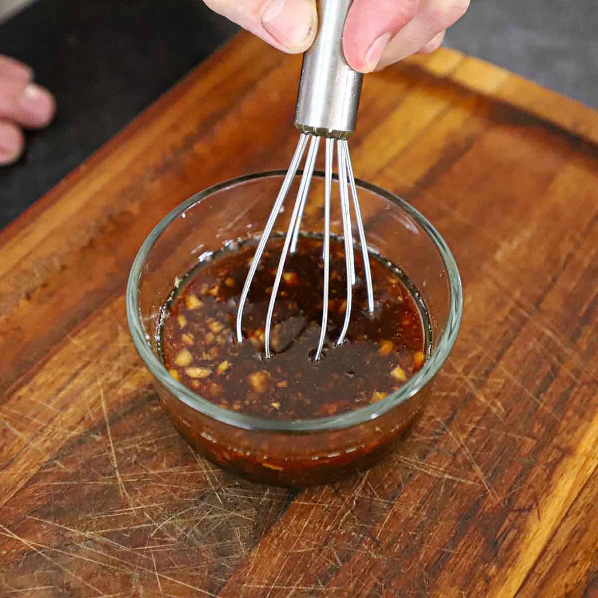 A person whisking a stir-fry sauce in a small glass bowl on a wooden cutting board.