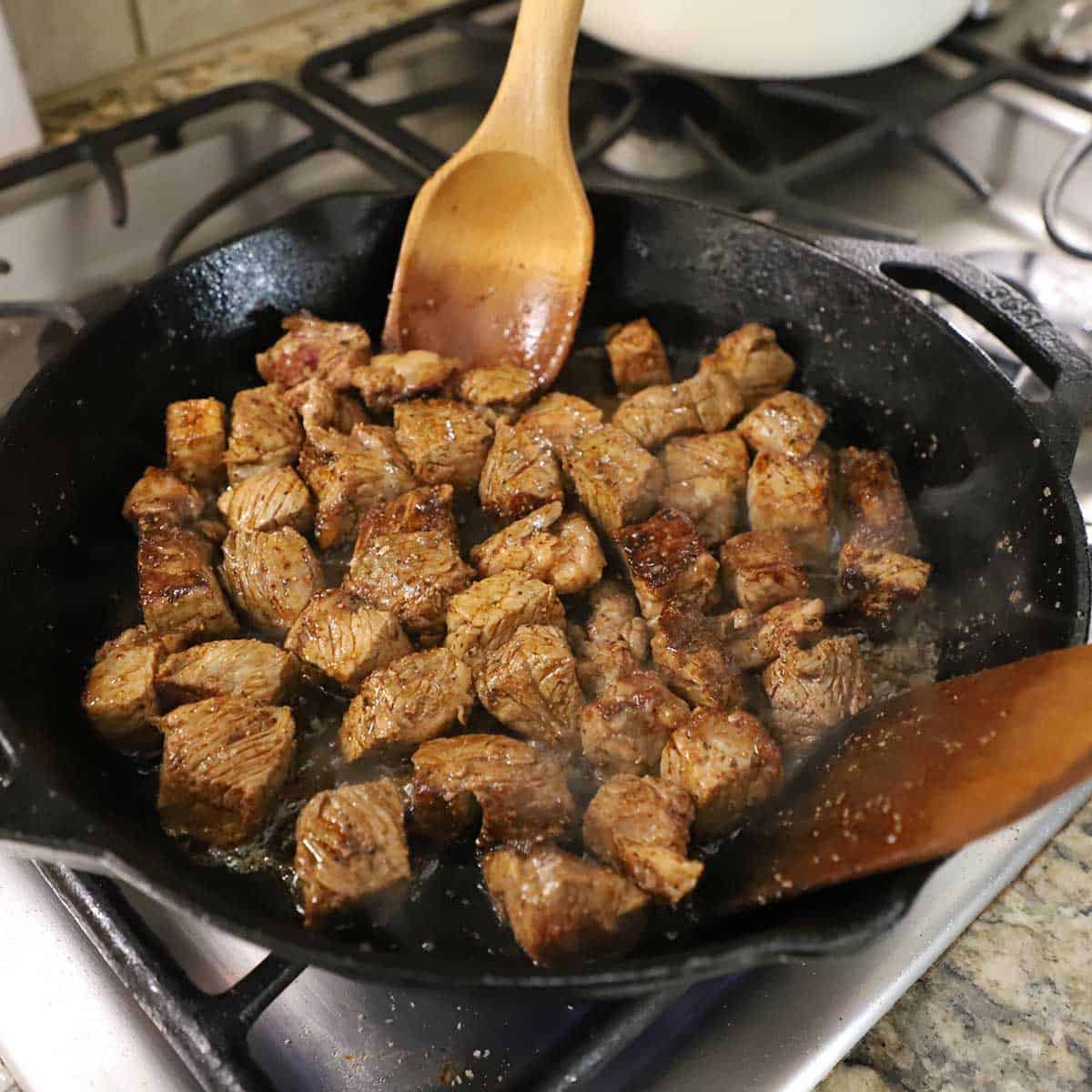 A person using two large wooden utensils to stir seared steak bites in a large cast-iron skillet on a gas stove.