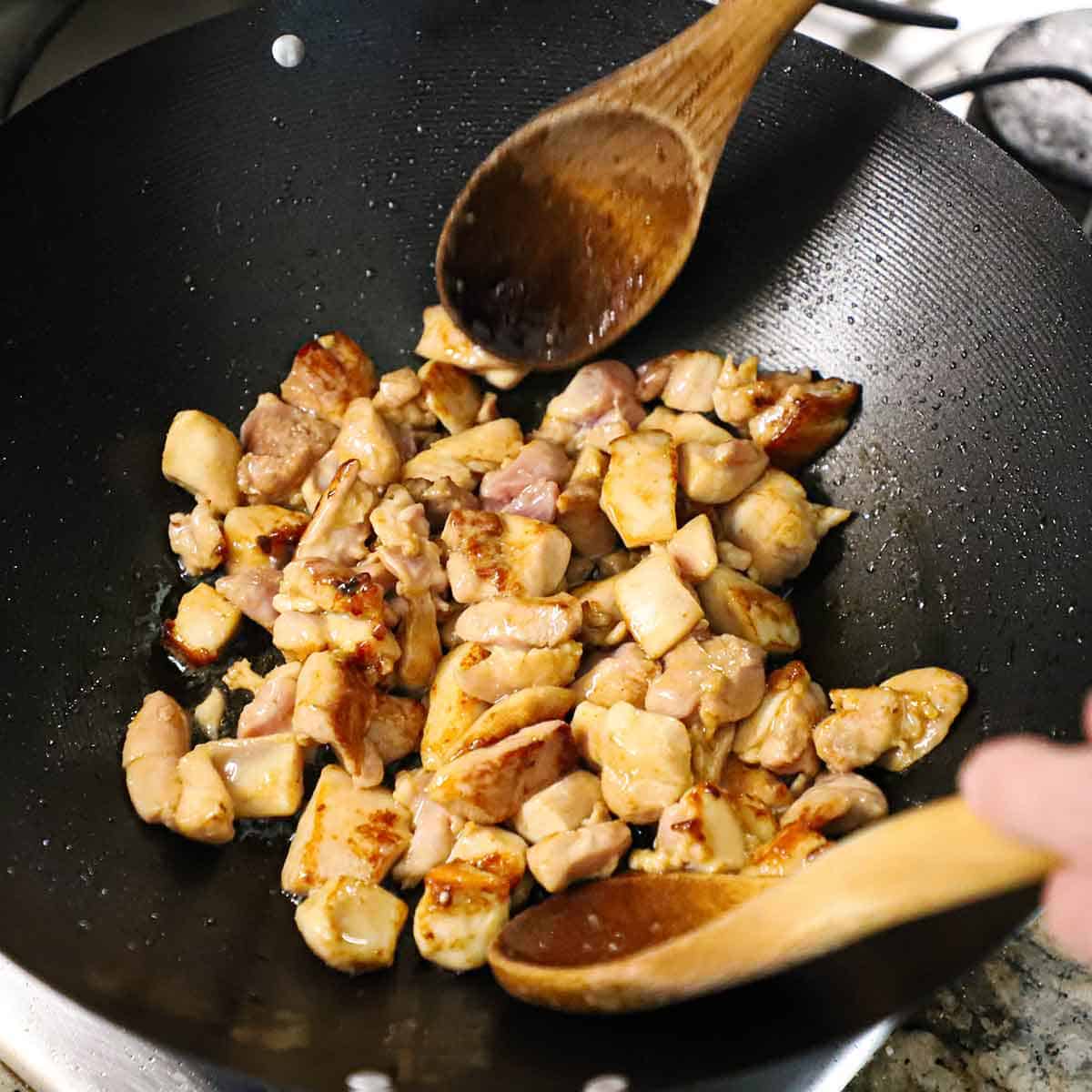 A person using two large wooden spoons to toss chunks of marinated chicken thighs that are being seared and cooked in a large wok on a gas stove.