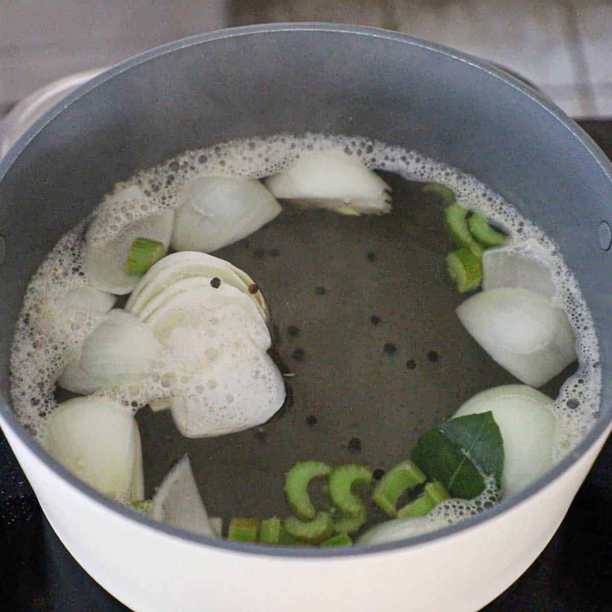 A large pot filled with simmering water and quartered onions, bay leaves, celery, and black peppercorns.